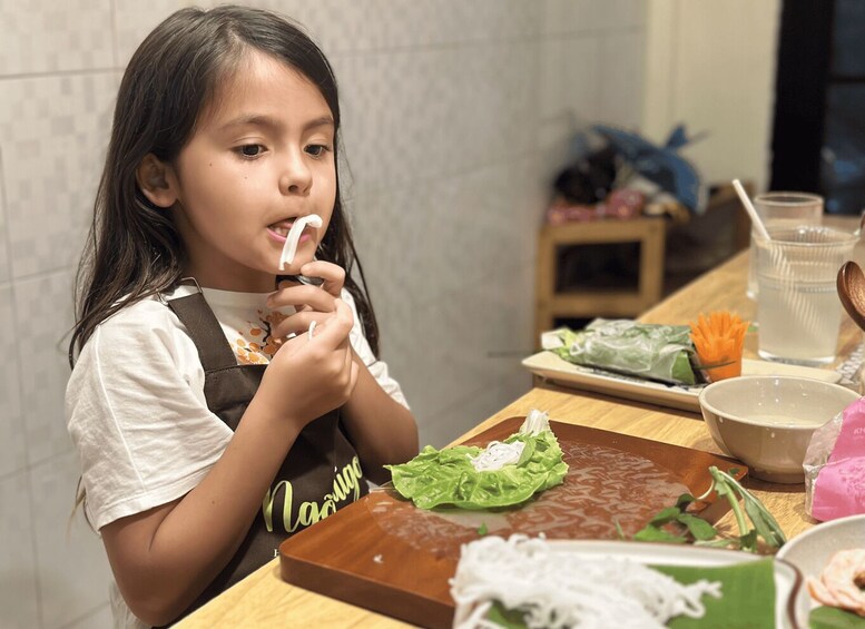 A child eating with a spoon, wearing a vest with 'Nasuya' on it, at a wooden table with food items and a drink. 4