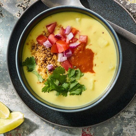 A bowl of soup garnished with diced tomatoes, red onions, cilantro, and a drizzle of sauce, with a lime wedge beside it. 3