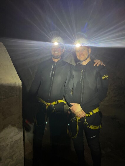Two people wearing helmets and harnesses, standing in a dimly lit cave. 2
