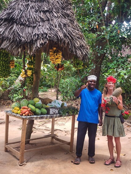 A man and a woman standing in a tropical setting with a thatched-roof structure and a table of fruits and vegetables. 2