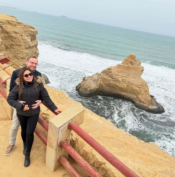 A couple standing on a sandy beach with a rocky outcrop in the background and the ocean waves crashing nearby. 3