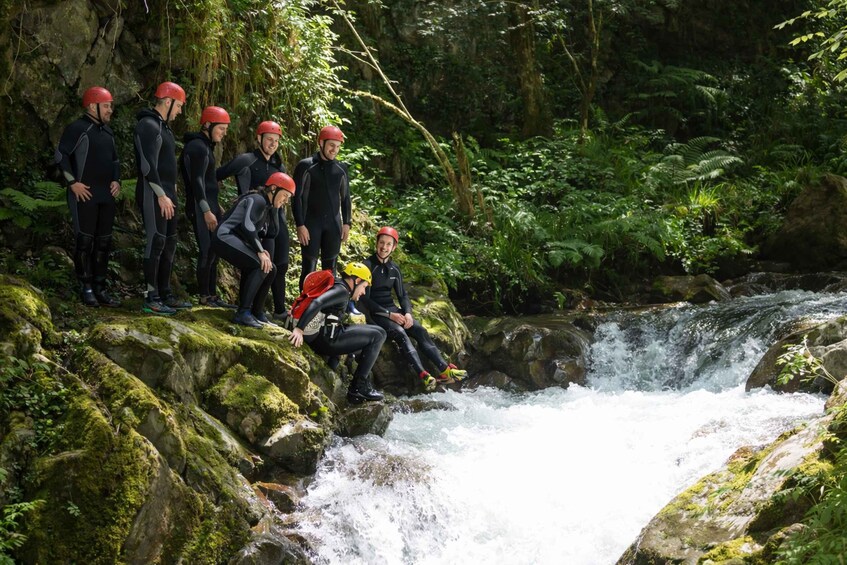A group of people in wetsuits and helmets sitting on rocks by a flowing river in a forested area. 3
