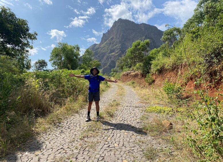 A person standing on a cobblestone path with arms outstretched, surrounded by greenery and a mountain in the background. 2