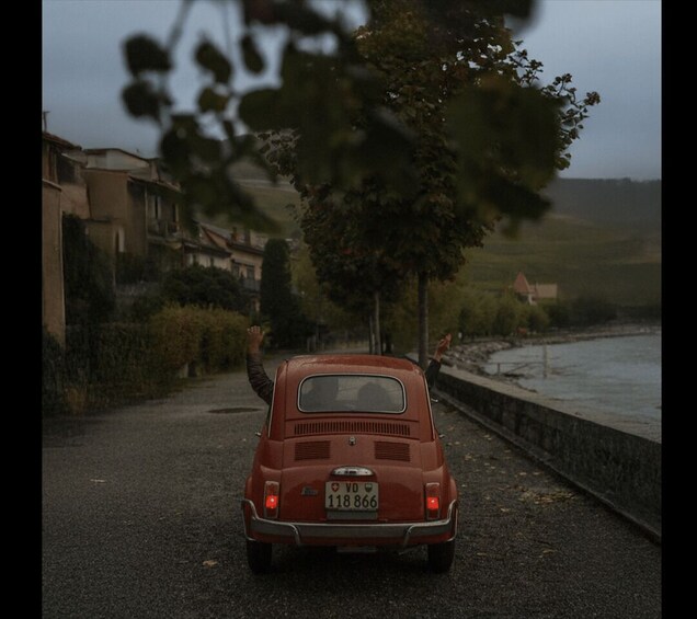 A vintage red car parked on a road with a river on one side and houses in the background. 4