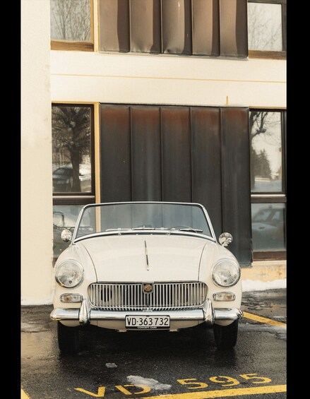 A vintage white convertible car parked in front of a building with large windows and a garage door. 2