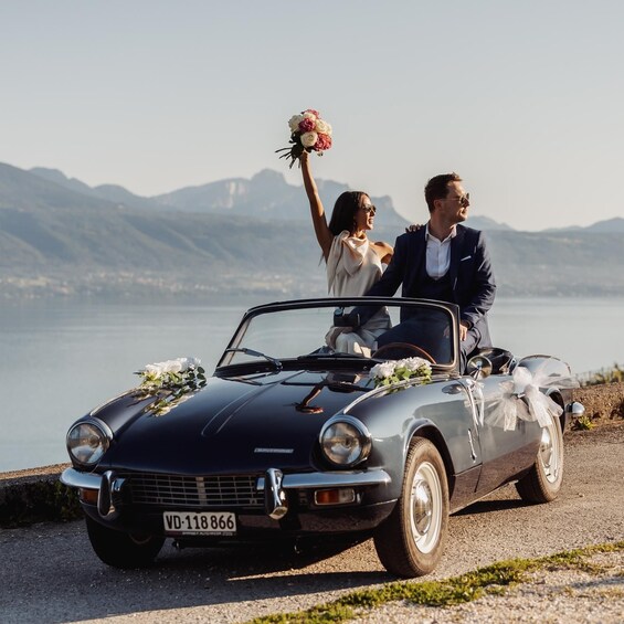 A vintage convertible car with a license plate, parked by a lakeside with mountains in the background. 1