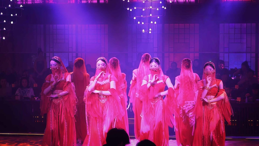 A group of dancers in red costumes performing on a stage with a purple-lit audience. 1