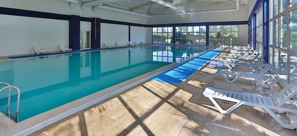 An indoor swimming pool with clear blue water, sunlit by large windows, and equipped with lounge chairs along the sides. 2