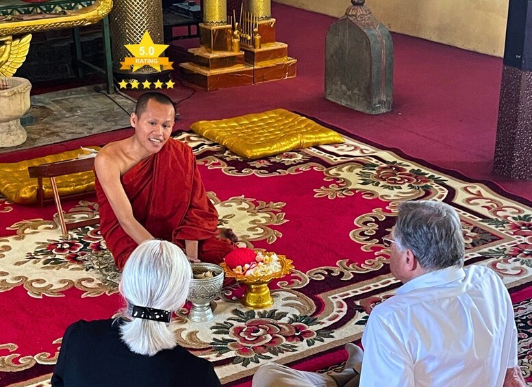 A monk in red robe sitting on a patterned carpet, with a bowl of offerings and a golden statue in the background. 4