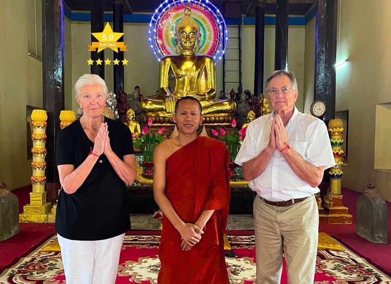A temple interior with a golden Buddha statue, a clock, and three people in prayer. 1