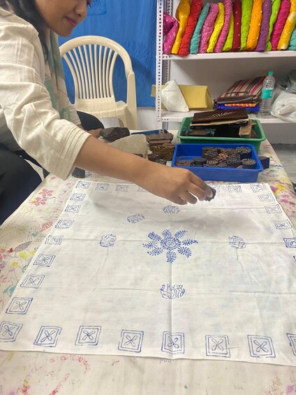 A person working on a fabric with a floral pattern, a blue tray with chocolate pieces, and colorful fabric rolls on a shelf. 2