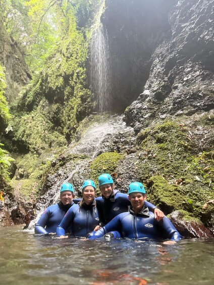 Four people in blue wetsuits and helmets sitting in a stream with a waterfall in the background. 3