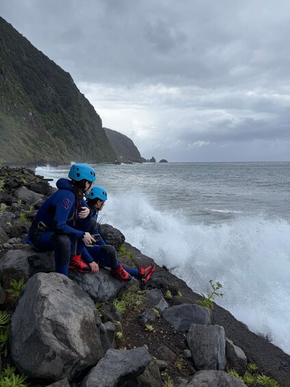 Two people in blue helmets sitting on rocks by the ocean, with waves crashing in the background. 4