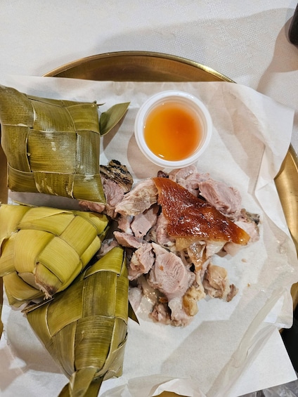 A plate of sliced meat, possibly pork, with a side of sauce and some food wrapped in leaves. 2