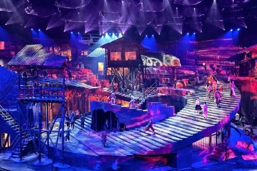 A stage with a wooden platform, audience seating, and a backdrop with a mountainous design. 1