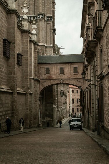 A historic stone archway in a narrow street with pedestrians and a parked car. 2