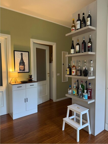 A room with wooden flooring, white cabinets, a small stool, and shelves with various bottles. 3