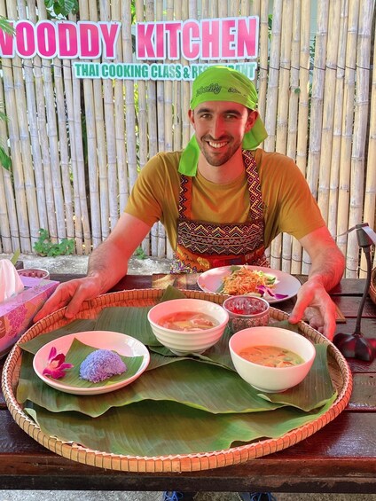 A person presenting a variety of Thai dishes on a woven tray, with a bamboo backdrop and a sign reading 'WOODY KITCHEN THAI COOKING CLASS & RESTAURANT'. 5
