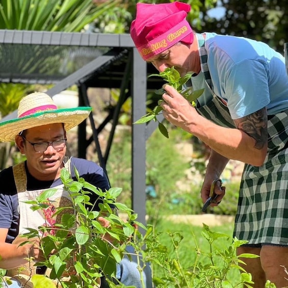 Two people gardening, one wearing a straw hat and apron, and the other in a blue shirt and checkered shorts. 1