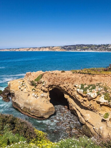 A coastal landscape with a prominent rock formation featuring a natural arch, a clear blue sea, and a distant shoreline with buildings. 1