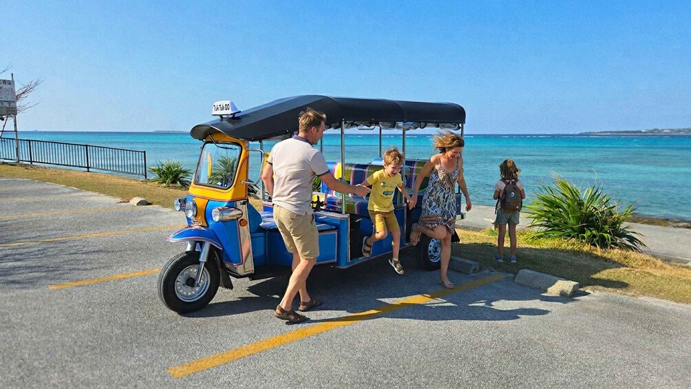 A blue and yellow tuk-tuk parked by the seaside with people boarding. 5