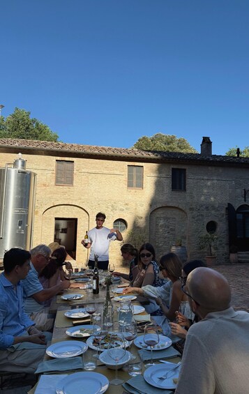 A group of people dining outdoors at a rustic table with a brick building in the background. 1