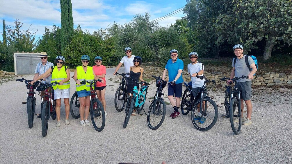 A group of people with bicycles in a gravel area with trees and a stone wall in the background. 2