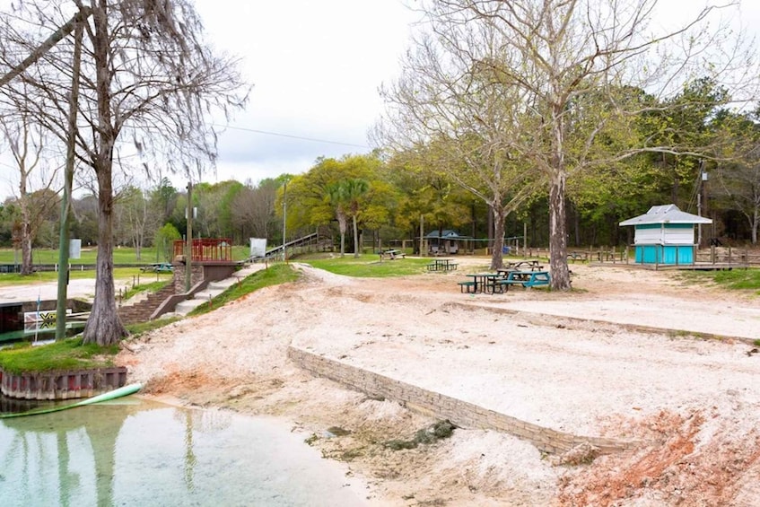 A park with picnic tables, a small blue shelter, and a sandy area near a water body. 2
