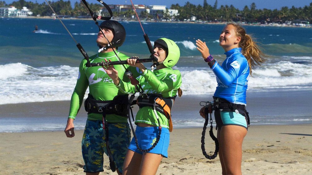 Three people on a beach with surfboards, wearing life vests and helmets, and holding kiteboarding equipment. 2