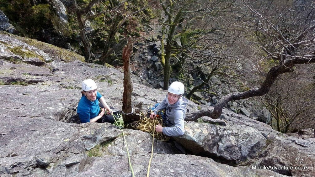 Two people rappelling on a rocky terrain with trees and moss in the background. 1