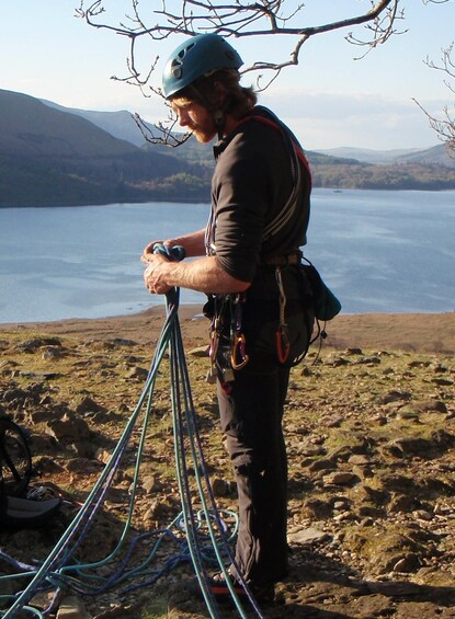 A person with climbing gear standing on rocky terrain near a body of water with mountains in the background. 3