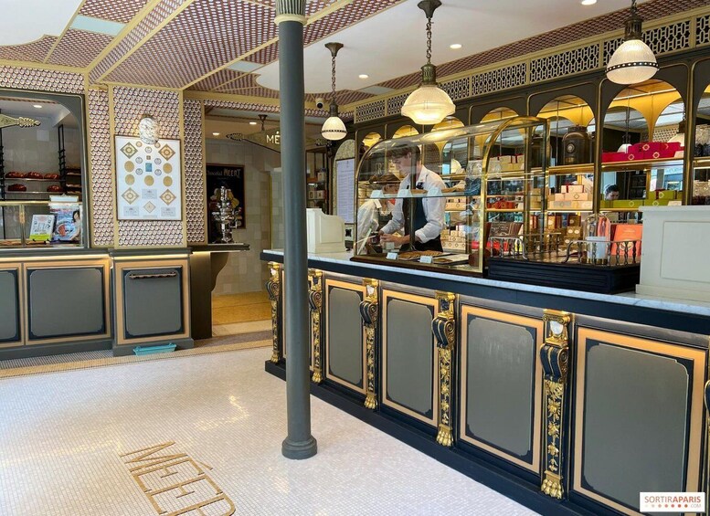 An interior with ornate gold detailing, a counter with a person behind it, and a patterned ceiling. 3