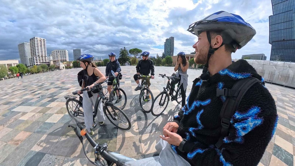 A group of cyclists on a paved area with buildings in the background. 1