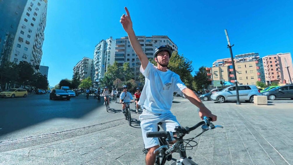 A person on a bicycle with a raised arm, other cyclists in the background, urban buildings, and parked cars. 3