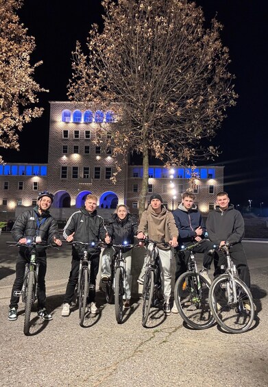 A group of people on bicycles in front of a building with illuminated blue windows. 2