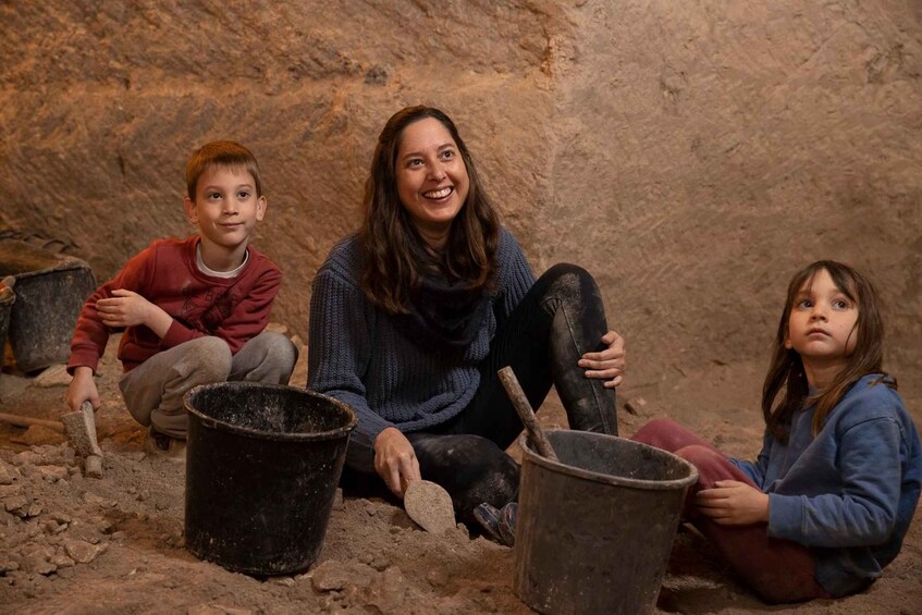 Three individuals sitting on the ground with buckets and shovels, surrounded by a rocky excavation site. 1