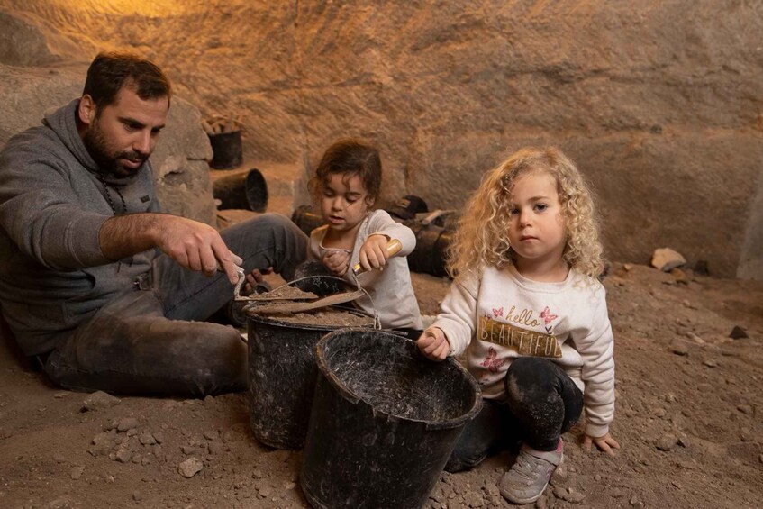 Two individuals sitting on the ground in a cave-like environment with dirt and rocks, surrounded by black buckets. 5