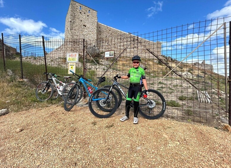 A person standing next to a mountain bike, with a historic stone tower and a fenced area in the background. 3
