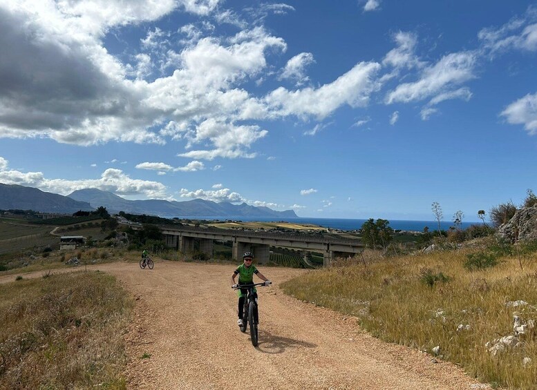 A gravel path with cyclists, a bridge, and a mountainous landscape under a partly cloudy sky. 1