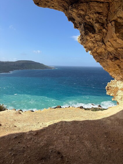 A coastal landscape with a rocky overhang, clear blue sea, and distant hills. 1