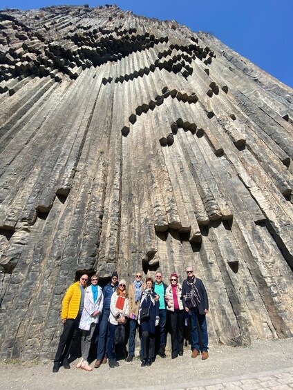 A group of people standing in front of a large, textured rock formation with distinct horizontal layers and hexagonal openings. 2