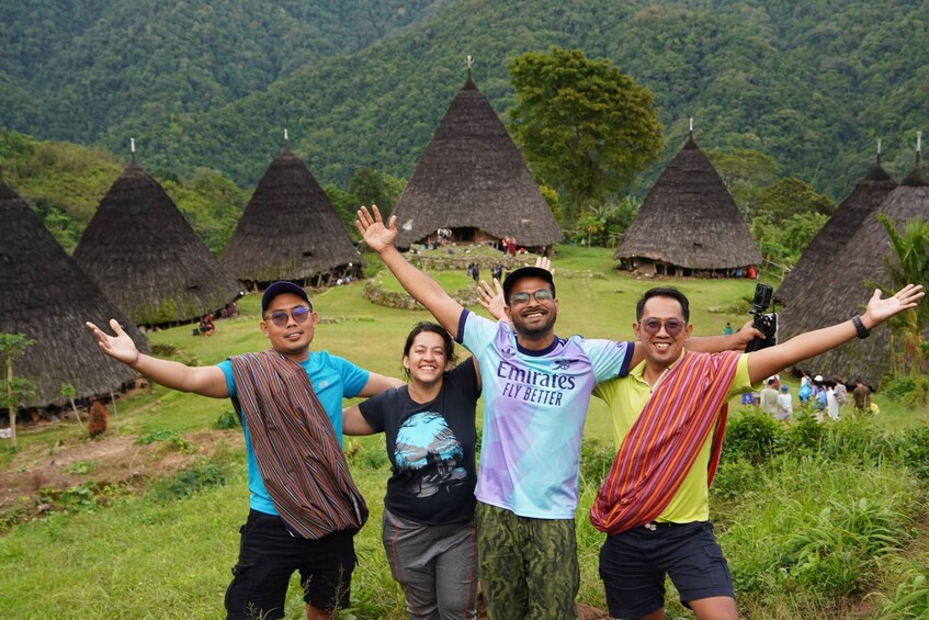 Four people standing in front of traditional conical huts in a grassy area with a mountainous backdrop. 1
