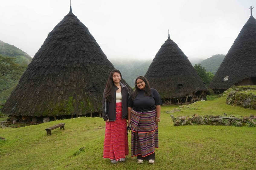 Two individuals standing in front of traditional conical huts with thatched roofs, set in a grassy area with a bench and a stone wall in the background. 3