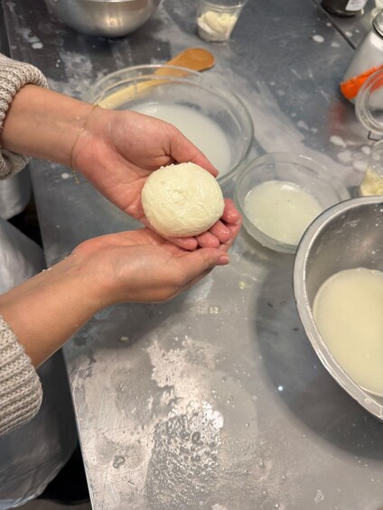 A person holding a round, white object, likely dough, with a bowl of white liquid in the background. 3