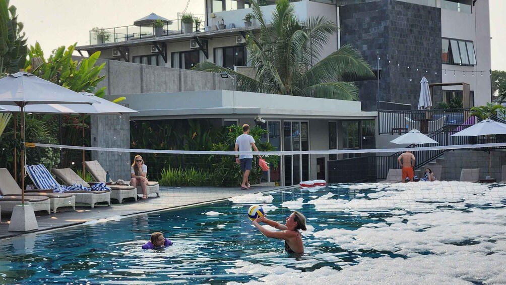 A swimming pool with people playing volleyball, lounge chairs, and a modern building in the background. 1