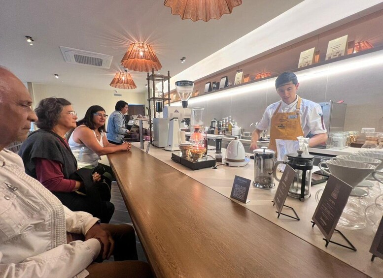 A coffee shop interior with patrons seated, a barista preparing drinks, and various coffee-making equipment. 1