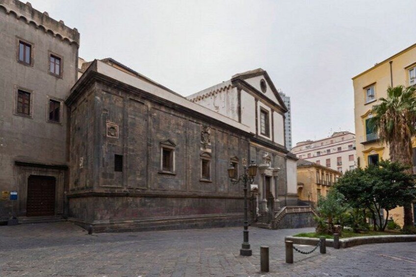 A historic building with a stone facade, a central entrance, and a small garden area with a palm tree. 2