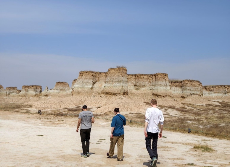 Three people walking on a sandy terrain with layered rock formations in the background. 1