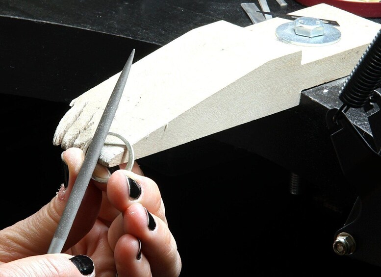 A hand holding a metal tool, possibly a file, against a white object secured in a vise. 2