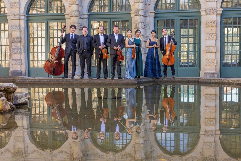 A group of musicians in formal attire standing in front of a classical building with a reflective water feature. 2